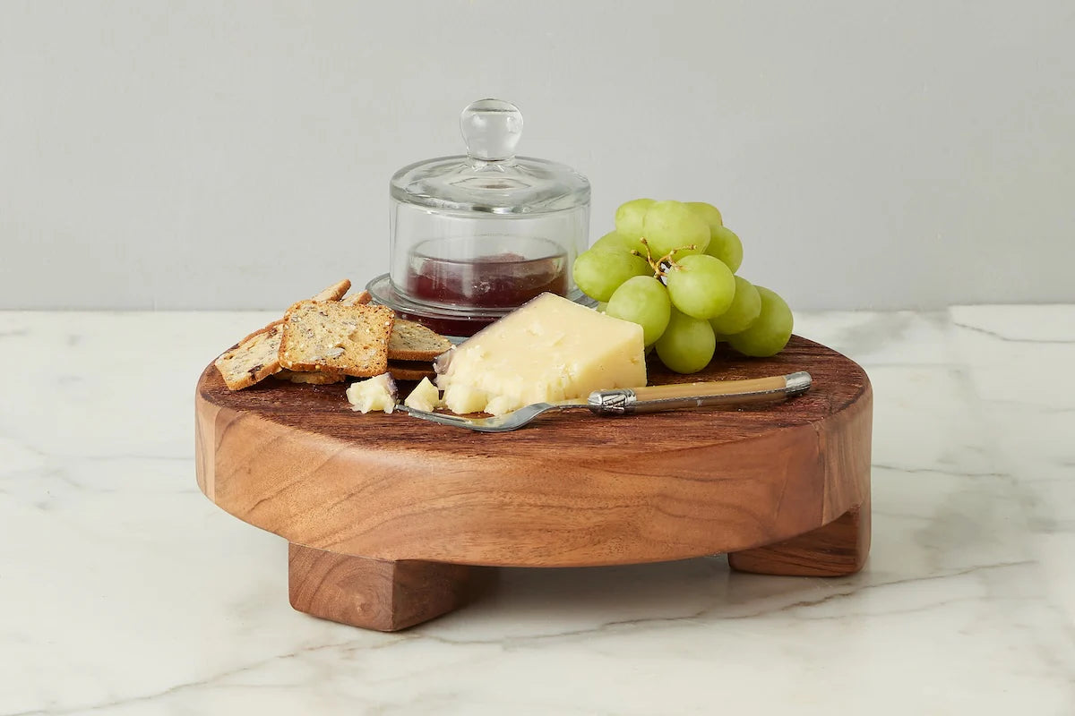 Wooden cheese board with cheese, grapes, round glass jam container, and bread on a marble surface.