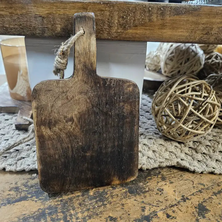 A dark brown wooden square chopping board with a rope attached to its top left corner, displayed on a table with a decorative background.
