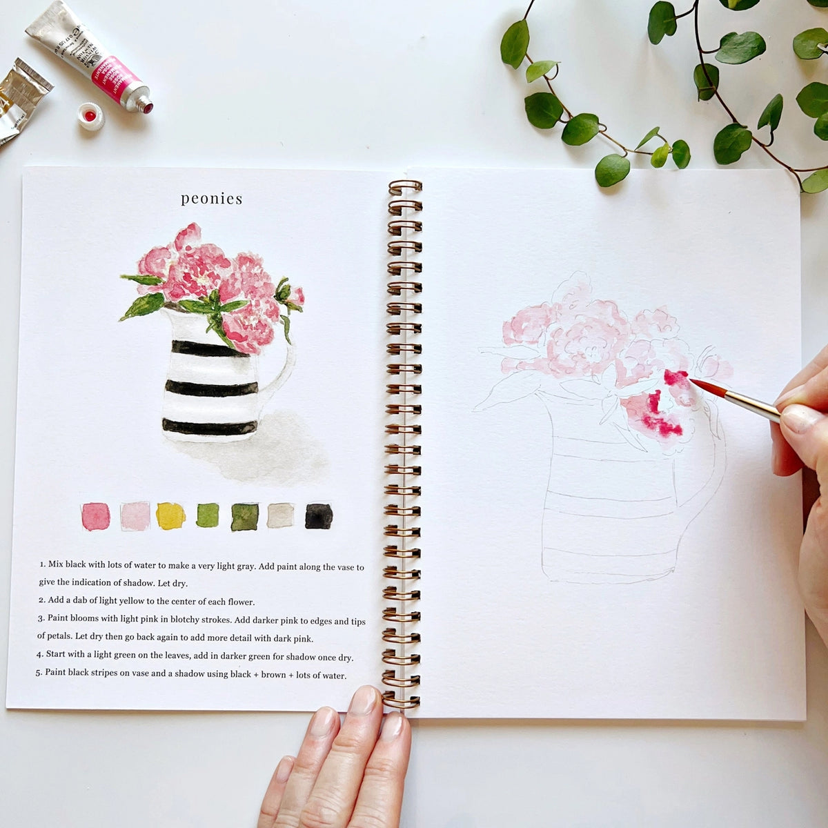 Open book with watercolor flowers and instructions with hand holding a paintbrush, on a white surface.