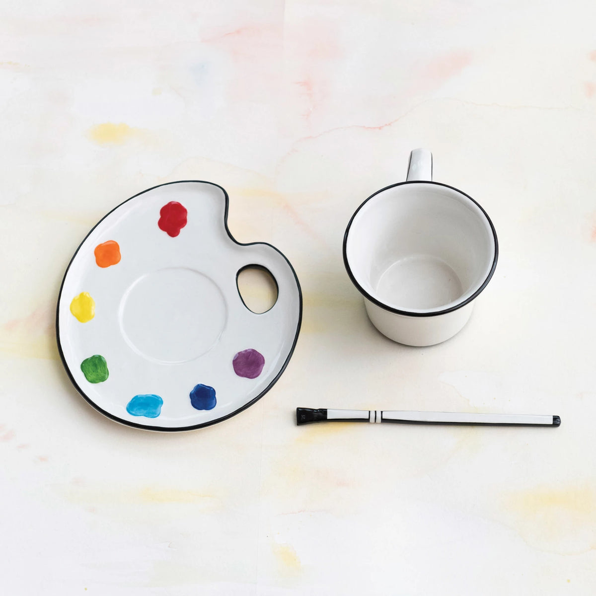 Paint palette shaped saucer with colorful paint swatches, a mug, and a brush shaped stirrer on a light background.