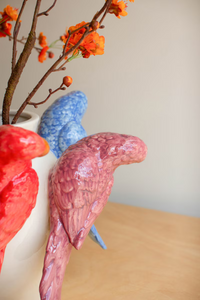 Three ceramic parrots in red, blue, and pink on a wooden surface with a neutral background