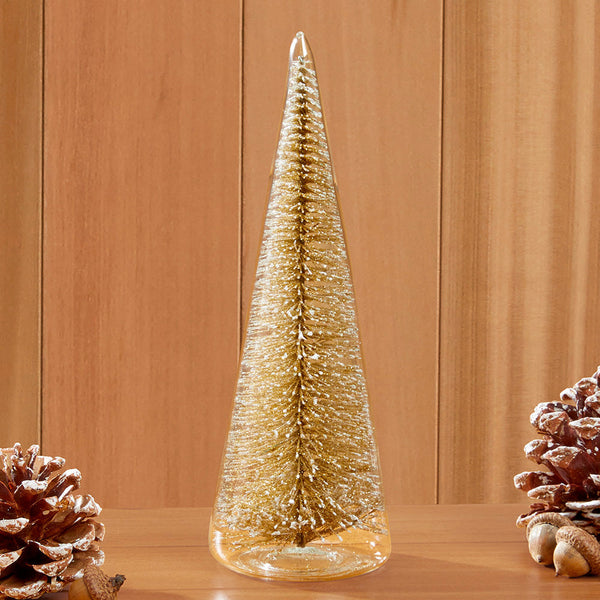 A tall, slender gold glitter Christmas tree decoration inside of glass, standing on a wooden surface, with pinecones on both sides against a wood-paneled background.