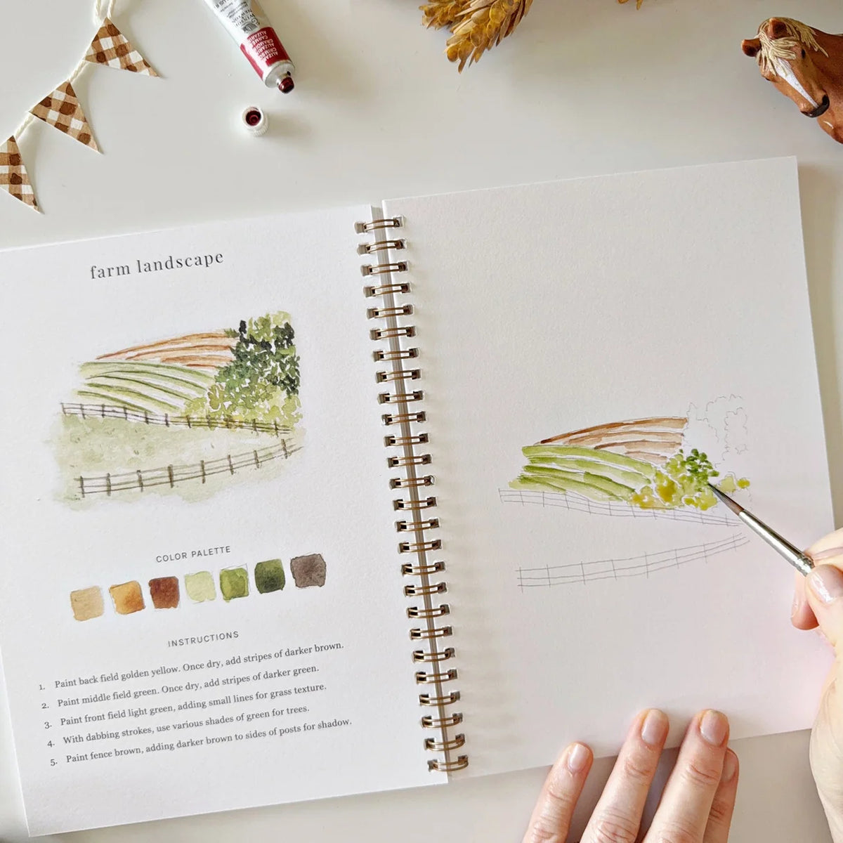 Artistic watercolor workbook with a person painting a farm landscape and color palette on a light surface.