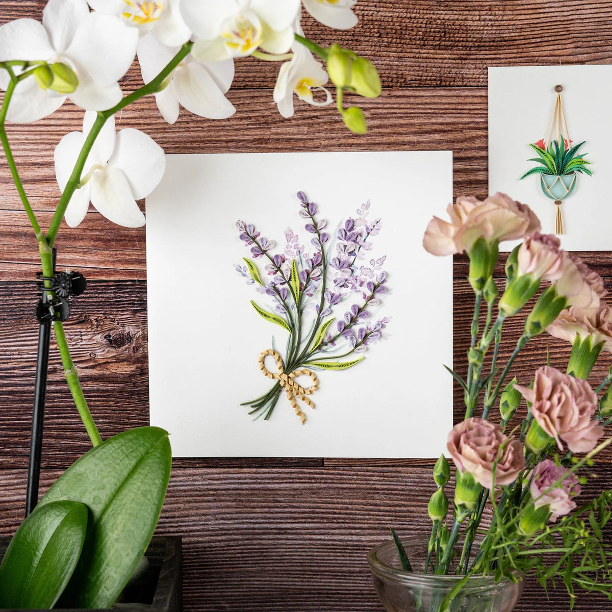 Floral arrangement with white orchids and pink flowers on a wooden surface, featuring a lavender quilled greeting card in the center.