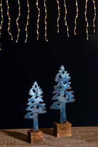 Two blue decorative trees on wooden stands with string lights in the background.