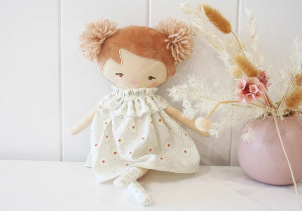 A fabric doll with brown hair in pom-poms, wearing a white dress with small orange dots, styled next to a pink vase with dried flowers on a white surface.