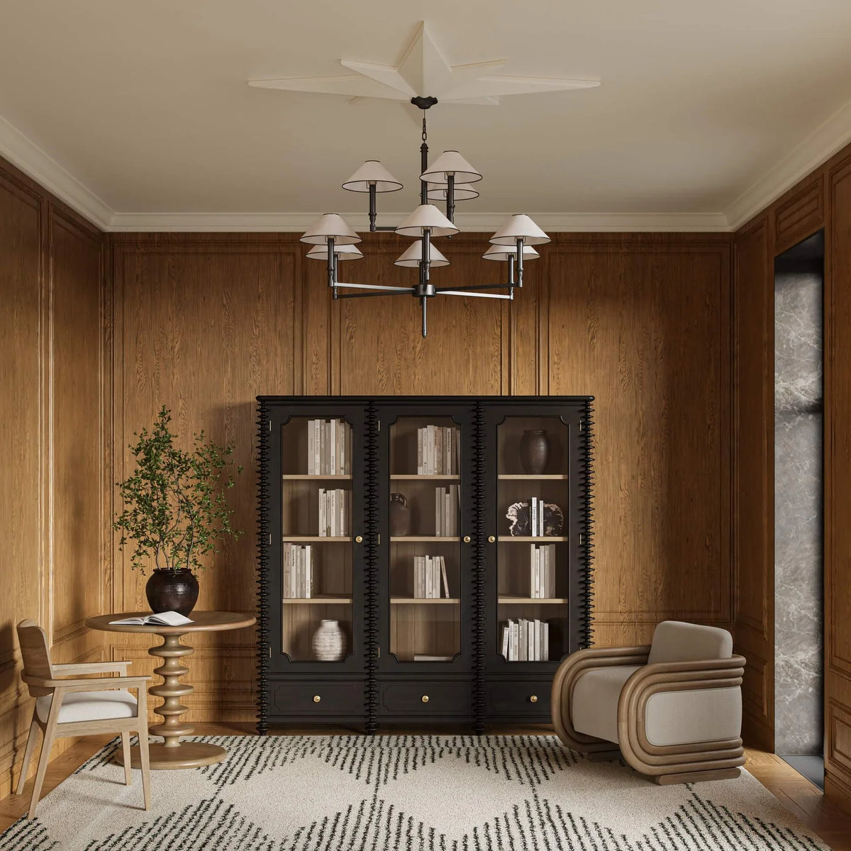 A room with wooden panel walls, two chairs, ad black and white rug, shelving unit with books, and an accent table with a plant on top of it.