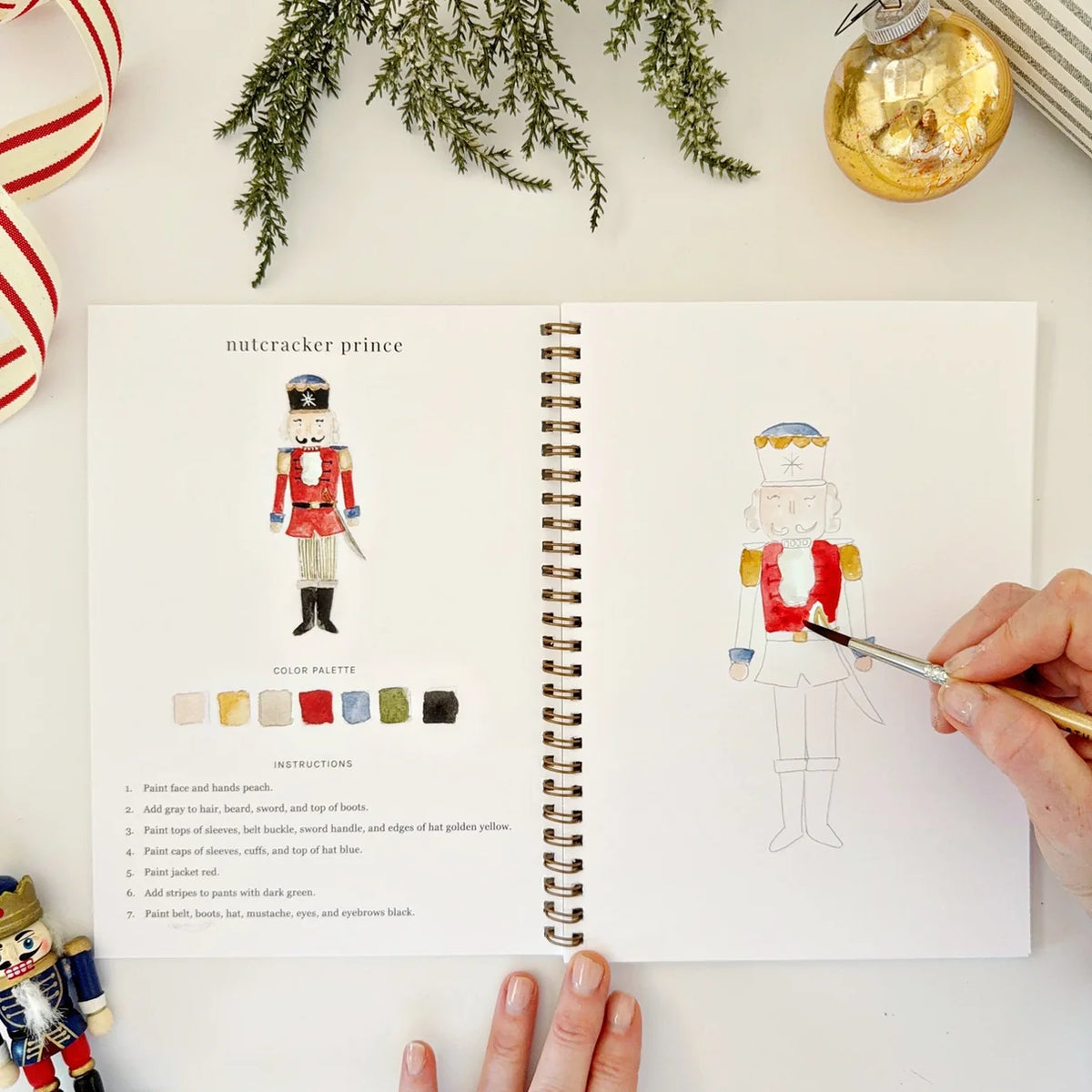 Person coloring a nutcracker outline with watercolor in a notebook with holiday themed objects around on the table.