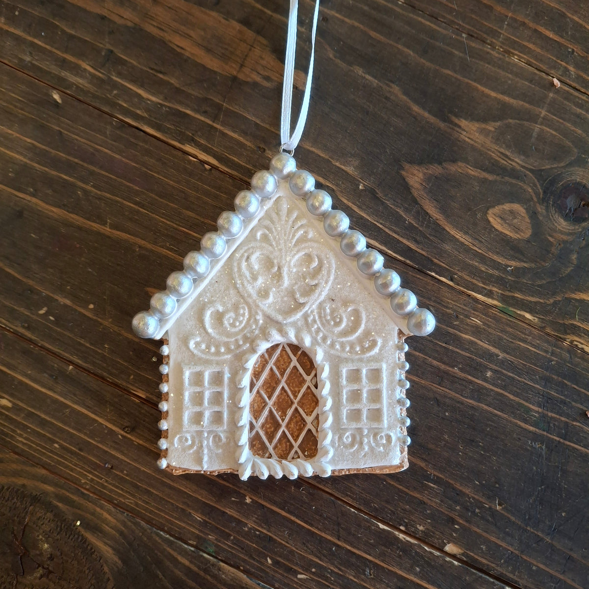 A white-painted gingerbread house ornament hanging from a white string against a dark brown wooden tabletop background.