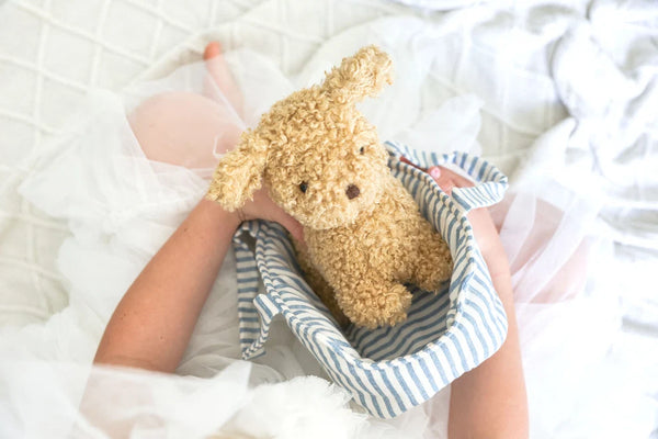 Person holding a small brown puppy dog toy in a striped bag on a white blanket