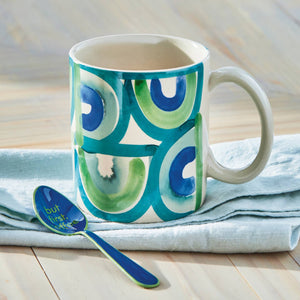 Mug with green and blue circular pattern on a light wooden surface with a spoon.