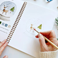 Person coloring a snow globe illustration in a notebook with a paintbrush.