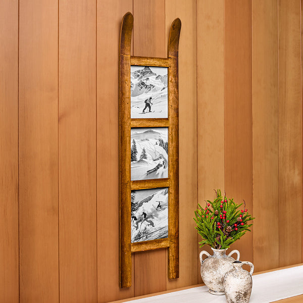 A rustic wooden ladder wall decor piece featuring three vertical panels with black-and-white ski and mountain illustrations, mounted on a warm wood-paneled wall beside two ceramic vases with greenery.